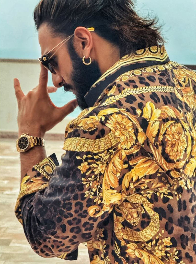 Ranveer is seen wearing a luxurious gold and black Versace shirt with intricate patterns, golden-rimmed black glasses, small golden hoops, and a classic Medusa Versace chain pendant set.