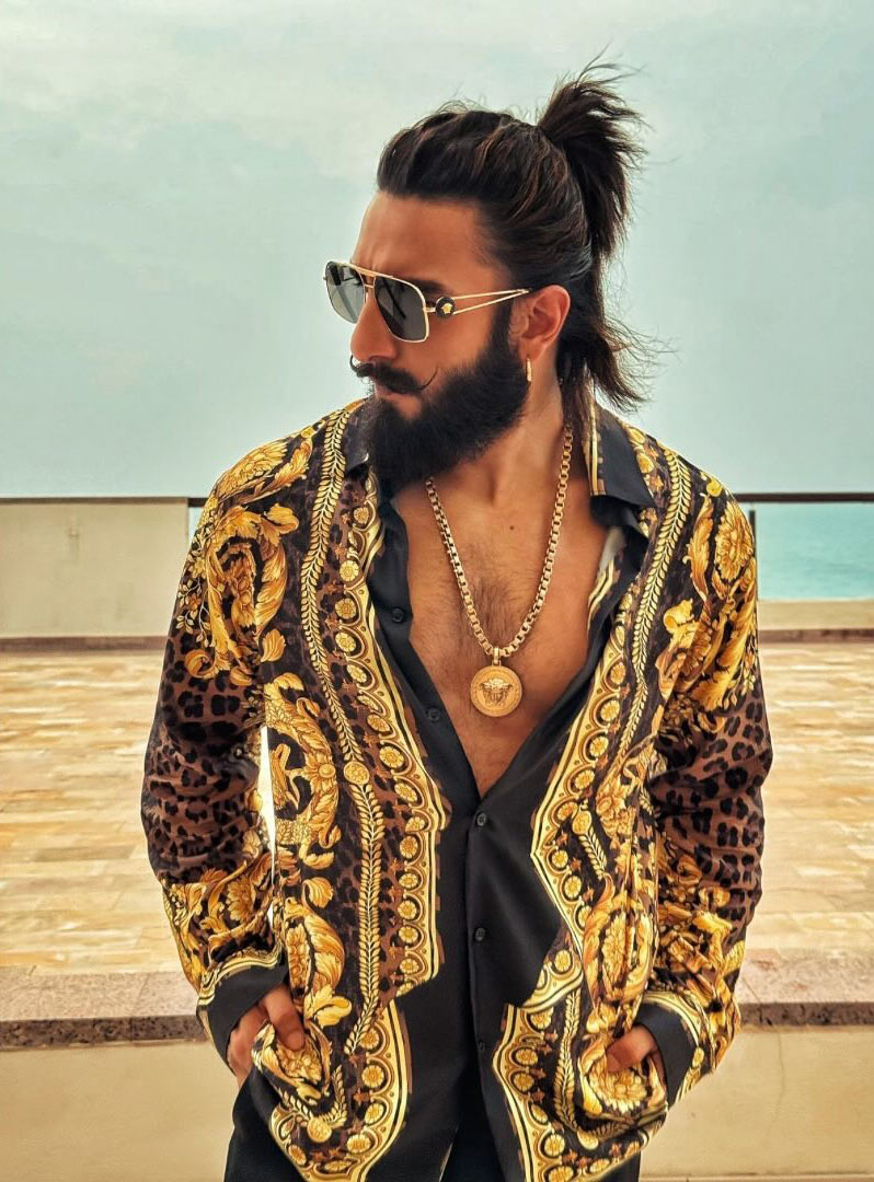Ranveer is seen wearing a luxurious gold and black Versace shirt with intricate patterns, golden-rimmed black glasses, small golden hoops, and a classic Medusa Versace chain pendant set.