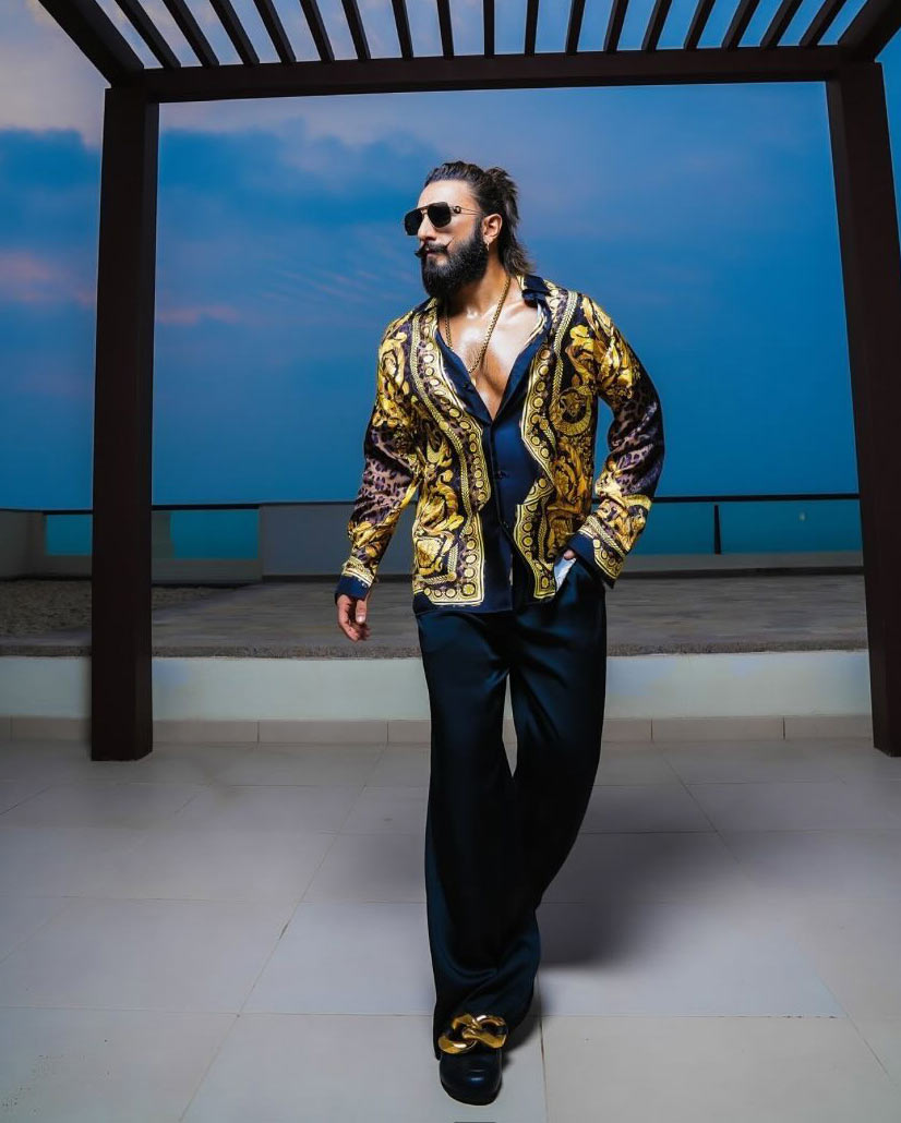 Ranveer is seen wearing a luxurious gold and black Versace shirt with intricate patterns, golden-rimmed black glasses, small golden hoops, and a classic Medusa Versace chain pendant set.