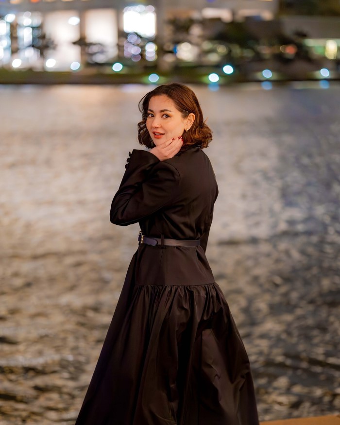 The photoshoot takes place by a serene waterfront at night, with blurred lights reflecting off the water, adding to the elegant ambiance.