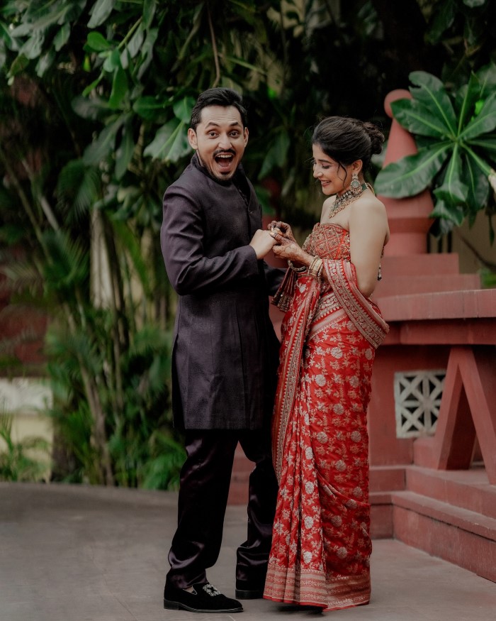 The bride-to-be stuns in a traditional red saree with intricate embroidery, paired with exquisite jewelry, while the groom looks dashing in a tailored black ethnic suit.