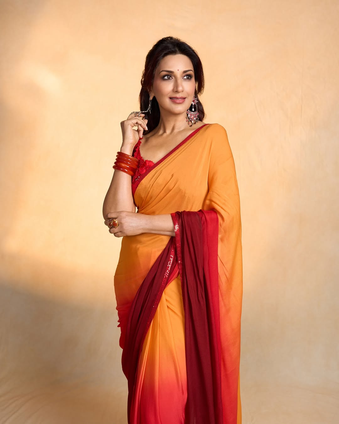 Sonali Bendre effortlessly blends tradition with a splash of contemporary flair in this vibrant orange and red saree–proof that age is just a number when you wear confidence and color with style and a hint of mischief.